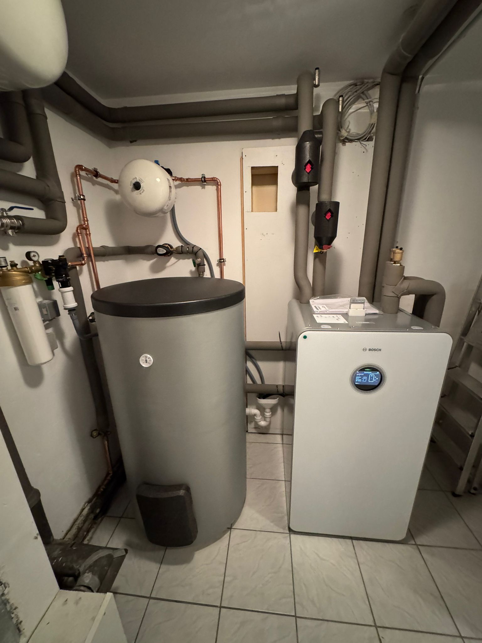 Eine Bosch-Wärmepumpe mit Speicher steht in einem gefliesten Heizungsraum, umgeben von gedämmten Leitungen und Armaturen.